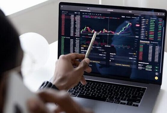A businessman using a smartphone while analyzing stock market graphs on a laptop indoors.